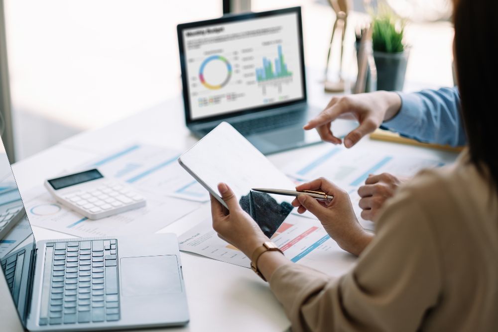 crm e automação rd station "Duas pessoas analisando dados em uma mesa de trabalho com notebooks, tablet e calculadora, representando a aplicação do CRM e da automação da RD Station na gestão e crescimento de lojas virtuais.
