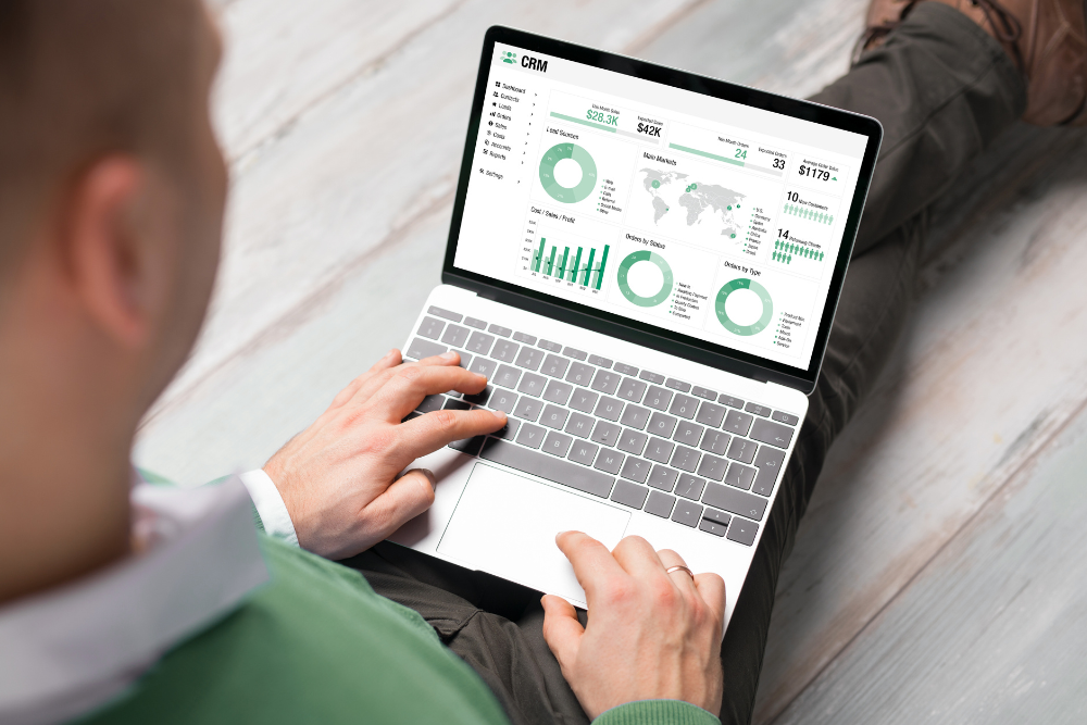 crm para e-commerce Homem sentado com notebook no colo, exibindo na tela diversos gráficos e painéis que representam um sistema de CRM para e-commerce, simbolizando análise de dados, automação e gestão de clientes.