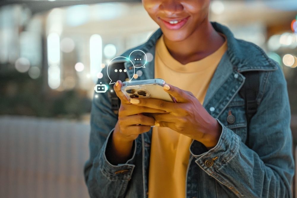 atendimento via chatbot Pessoa sorrindo enquanto utiliza o celular para se comunicar, com ícones digitais representando mensagens e robô, simbolizando o atendimento via chatbot.
