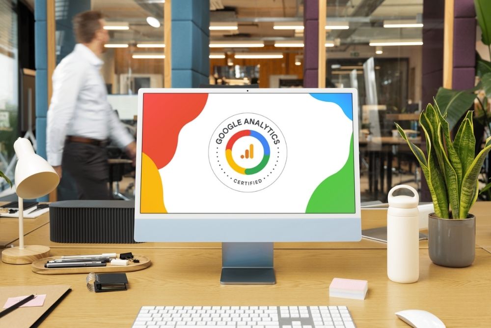 agência certificada GA4 Imagem de um escritório moderno com um computador central exibindo o selo "Google Analytics Certified". O ambiente é corporativo, com objetos de trabalho sobre a mesa, uma planta decorativa e uma pessoa desfocada ao fundo. A imagem representa profissionalismo e reforça a credibilidade de uma agência certificada GA4.
