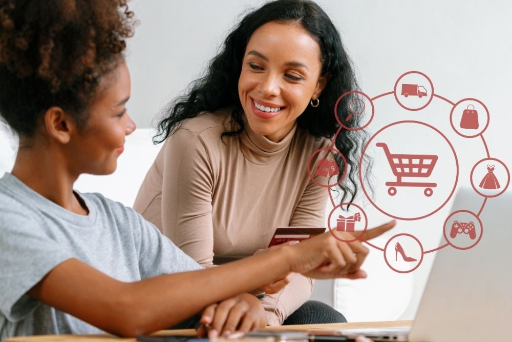 Tendências de comportamento de consumo Duas mulheres sorridentes analisam opções de compras online em um notebook, com ícones representando diferentes categorias de produtos. A imagem ilustra as Tendências de comportamento de consumo para o segundo semestre de 2025, destacando a digitalização, conveniência e variedade no e-commerce.