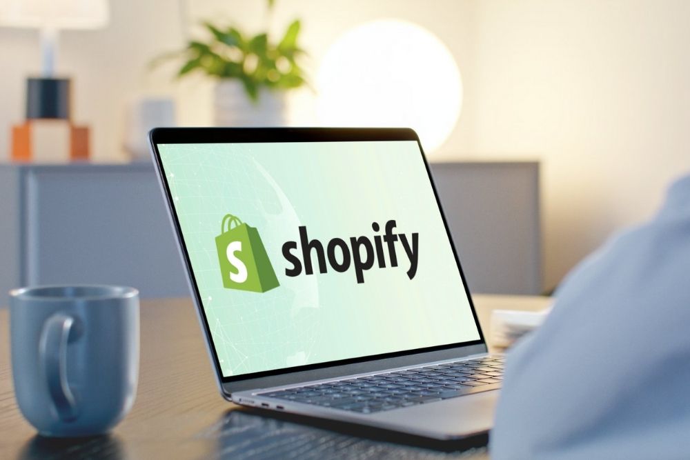 Cross border com Shopify Close de um notebook sobre uma mesa de trabalho exibindo o logo da Shopify, ilustrando um lojista planejando sua estratégia de vendas internacionais através do cross-border com Shopify.