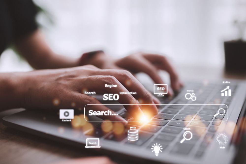 SEO para e-commerce em 2025 Mãos de uma pessoa digitando em um notebook, com sobreposição digital de palavras relacionadas a otimização de mecanismos de busca (SEO) e ícones representando elementos do SEO para e-commerce em 2025, como pesquisa, conteúdo e análise de dados.