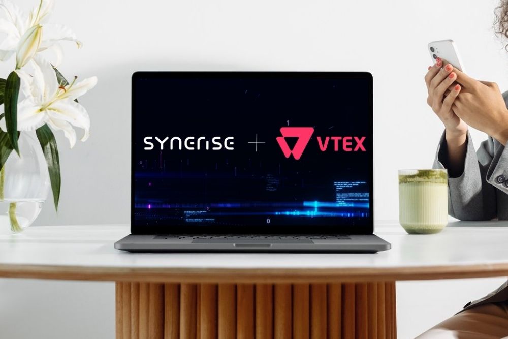 Synerise e VTEX Um notebook sobre uma mesa de madeira exibe na tela o logo da Synerise e da VTEX conectados pelo símbolo de "mais". Ao fundo, uma pessoa segura um celular e há um copo com uma bebida verde e flores brancas em um vaso.