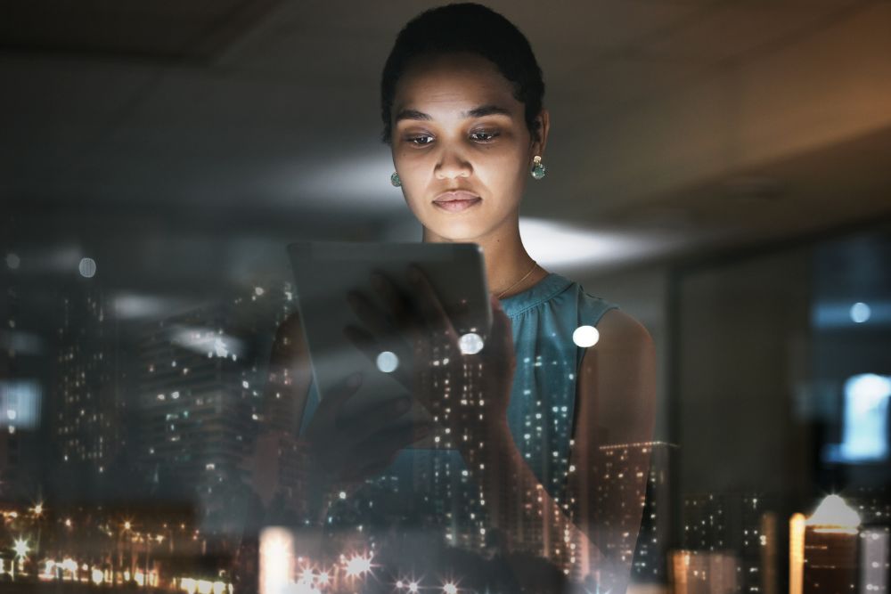 IA Corporativa Mulher afro-americana concentrada, olhando para um tablet em um ambiente de escritório com reflexos da cidade noturna, simbolizando a IA corporativa.
