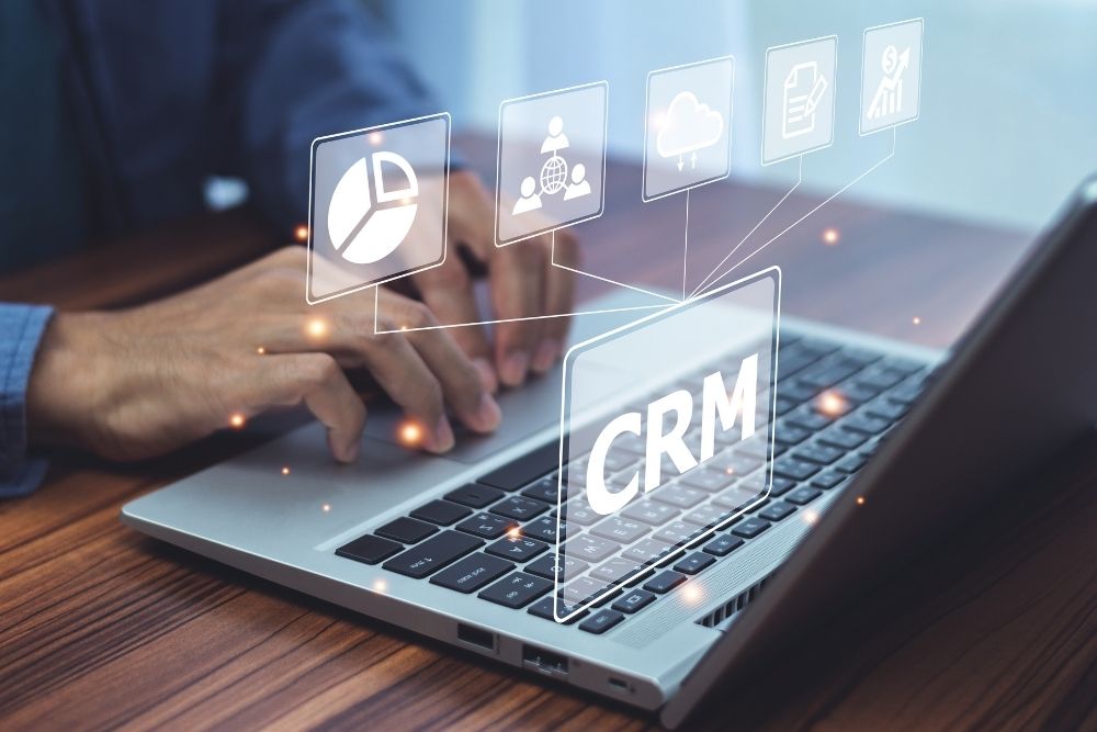 CRM para e-commerce Pessoa digitando em um notebook com ícones digitais flutuando relacionados a análise de dados, comunicação e automação, com destaque para o termo “CRM” ao centro, representando o uso de crm para e-commerce.