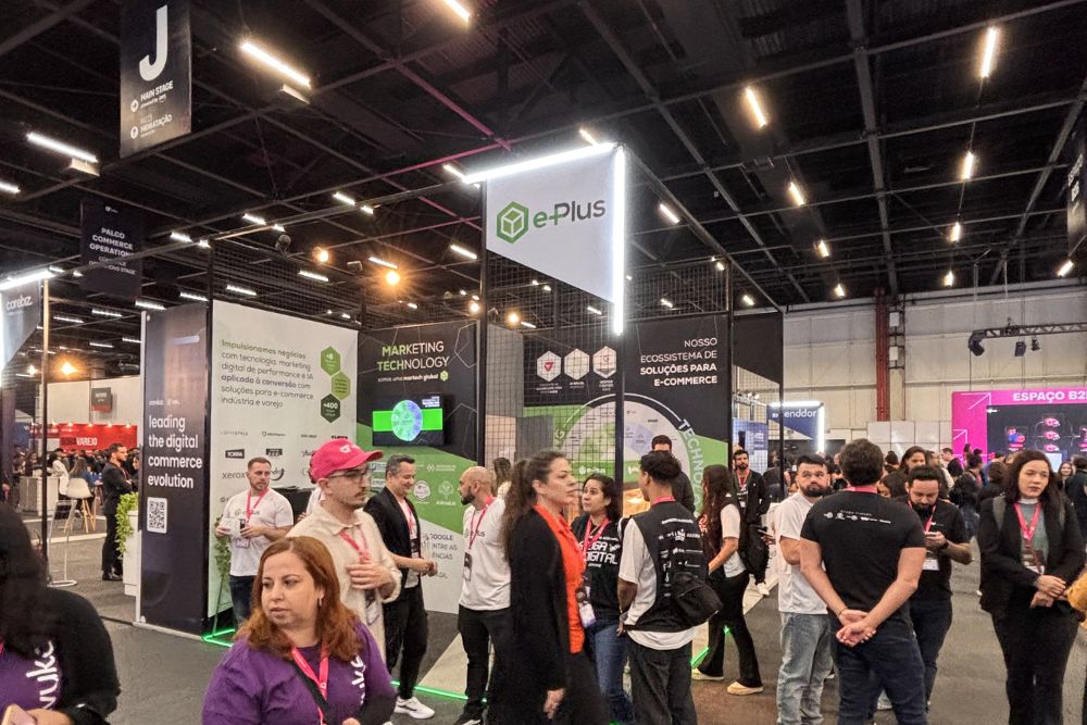 prepare-se para o VTEX DAY 2026 Fotografia panorâmica do movimentado estande da Agência e-Plus durante o vtex day 2026. O espaço moderno destaca-se no pavilhão com um grande letreiro luminoso suspenso contendo o logotipo da e-Plus, além de painéis informativos exibindo os termos 'Marketing Technology' e 'Nosso ecossistema de soluções para e-commerce'. Dezenas de profissionais, especialistas e visitantes interagem nos arredores do estande, que possui iluminação no piso em verde neon, refletindo o intenso ambiente de inovação e networking do evento.