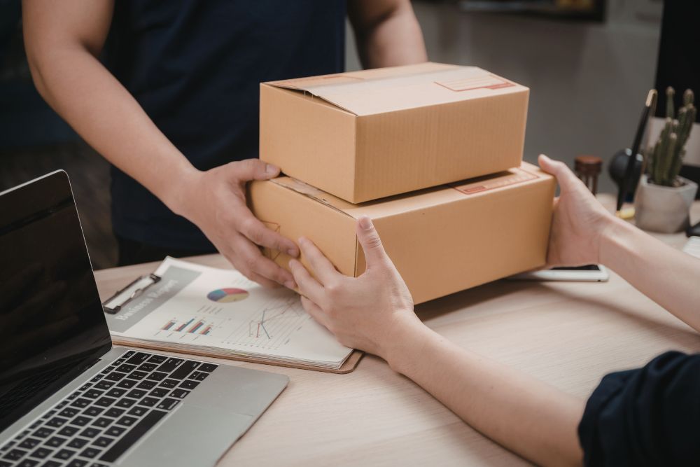 Frete caro Detalhe das mãos de duas pessoas entregando caixas de papelão sobre uma mesa com notebook e gráficos financeiros, ilustrando a operação logística de um e-commerce na busca por soluções para reduzir o impacto do frete caro nas vendas.