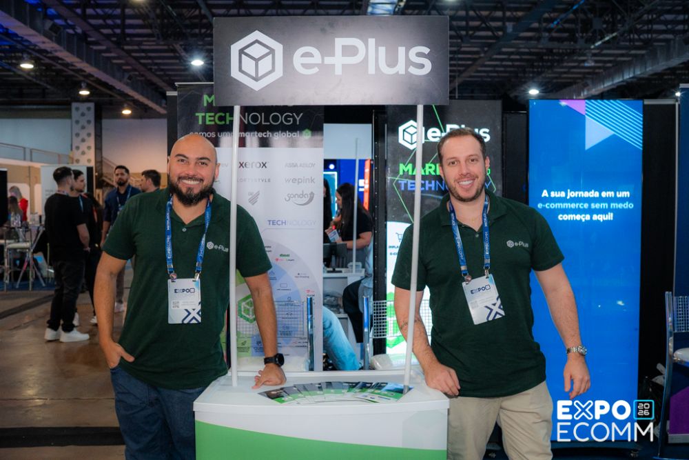 ExpoEcomm Blumenau Dois especialistas da agência e-Plus sorrindo no estande da empresa durante a expoecomm blumenau, com banners sobre marketing e tecnologia ao fundo.
