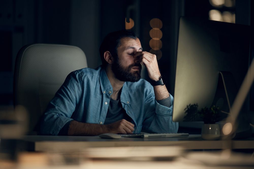 Problemas operacionais que limitam o seu e-commerce Homem com expressão de exaustão, esfregando o rosto em frente à tela do computador à noite, representando o estresse causado por problemas operacionais no e-commerce.