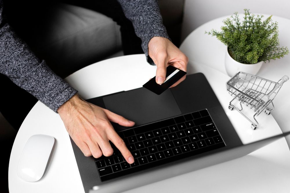 Mãos de um consumidor segurando um cartão de crédito enquanto digita em um notebook para finalizar uma compra via checkout transparente, com um mini carrinho de supermercado sobre a mesa branca ao lado.