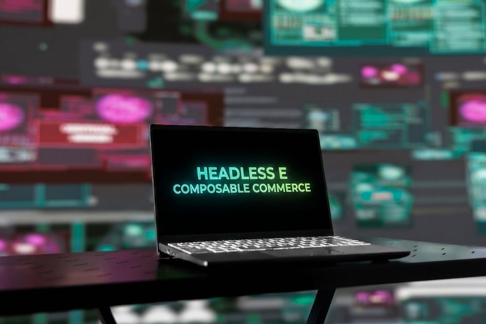 Notebook aberto sobre uma mesa escura exibindo na tela o texto Headless e Composable Commerce em letras luminosas verdes, com painéis digitais e gráficos de dados desfocados ao fundo.