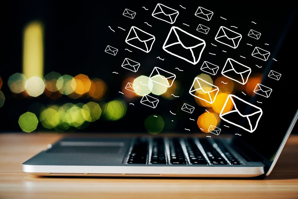 Notebook aberto sobre uma mesa de madeira à noite com ilustrações de envelopes brancos voando para fora da tela, representando envios de e-mail marketing, com luzes urbanas desfocadas ao fundo.