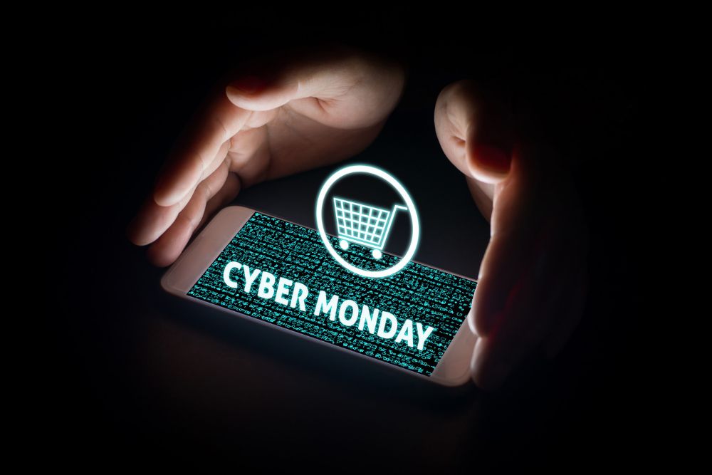 Mãos ao redor de um celular com a tela iluminada exibindo Cyber Monday e um ícone de carrinho de compras sobre um fundo de código digital